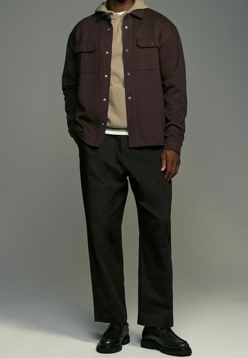 Homme portant une veste marron foncé boutonnée par-dessus un sweat à capuche beige, pantalon noir et chaussures noires, debout devant un fond gris uni.