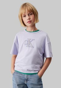 Camiseta de manga corta en color lila con logo bordado "CK", superpuesta sobre una camiseta verde, combinada con jeans de mezclilla azul claro. Textura de algodón suave.