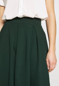 Kvinna som bär en höga midjan, mörkgrön plisserad kjol och en inskjuten, kortärmad vit blus, med höger arm avslappnad vid sidan.