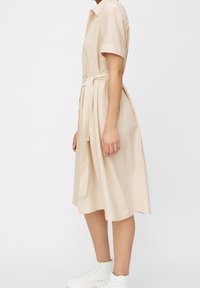 Abito color beige con colletto, maniche corte, un motivo a righe testurizzato, una cintura annodata in vita, che arriva fino a metà polpaccio, abbinato a scarpe da ginnastica bianche.