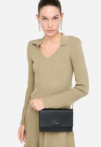 Borsa crossbody nera con texture, dotata di una tracolla a catena dorata, caratterizzata da un pattina pieghevole e logo del marchio impresso. Il modello indossa un outfit verde a coste.