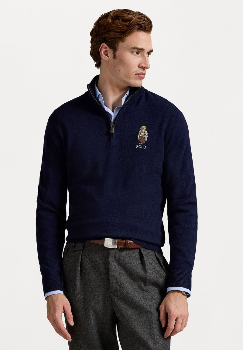Navy zip-up sweater with a high collar, featuring a small embroidered Polo logo and a detailed graphic of a character on the chest.