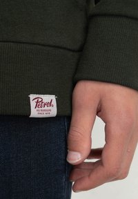 Ciemnozielona bluza z fakturą prążkowaną, z białą metką z napisem "Petrol" na czerwono, przyszytą u dołu nad dżinsowym materiałem.