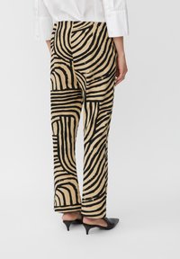 Pantalon en tissu beige avec de grosses rayures noires ondulées. Design évasé avec une texture lisse, porté avec des chaussures noires à talons bas.