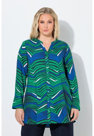 Kvinde iført en langærmet knap-op tunika med grønne, blå, sorte og hvide abstrakte bølgede striber, stående mod en ensartet lys baggrund.