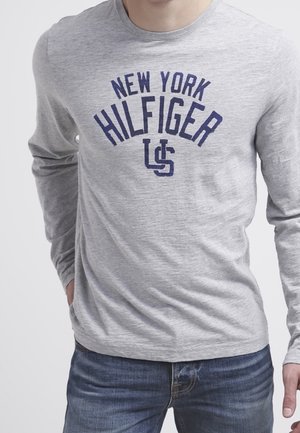 Helles graues Langarm-T-Shirt mit navyblauem Text "NEW YORK HILFIGER" in einer geschwungenen Schrift, kombiniert mit blauen Jeans.