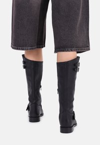 Botas de cuero negras hasta la rodilla con dos hebillas plateadas, cremalleras a los lados y punta redonda, combinadas con pantalones oscuros grises recortados.