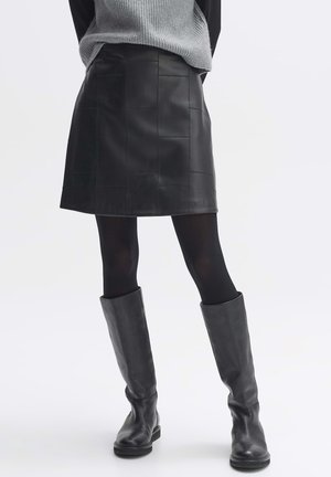 Woman wearing a black leather patchwork skirt, black tights, black knee-high leather boots, and a gray sweater, standing against a white background.