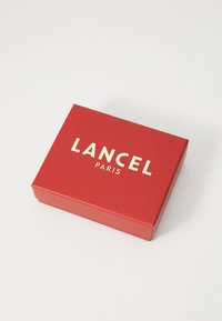 Czerwony prostokątny pudełko wykonane z papieru o gładkiej fakturze, z napisem "LANCEL" i "PARIS" w złotych literach na górze.