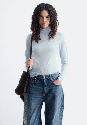 Femme aux cheveux bouclés portant un haut bleu clair à manches longues et un jean bleu tient un sac à bandoulière sombre avec une main dans la poche.