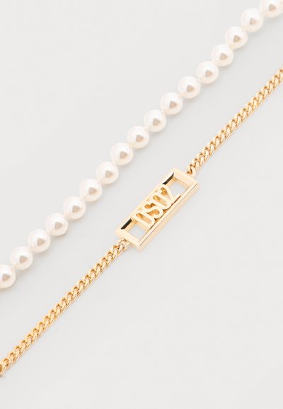 Βραχιόλι από χρυσή αλυσίδα με ορθογ ώνιο μενταγιόν «DSW» δίπλα σε σειρά στρογγυλών λευκών μαργαριταριών σε λευκό φόντο.