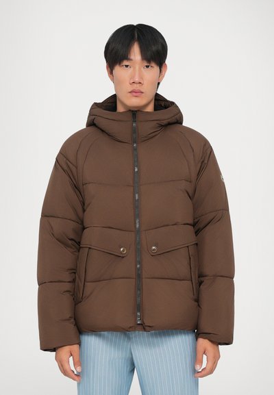 Giacca puffer marrone con cappuccio, dotata di una cerniera frontale, due tasche con bottone e una texture trapuntata. Indossata con pantaloni a righe blu chiaro.