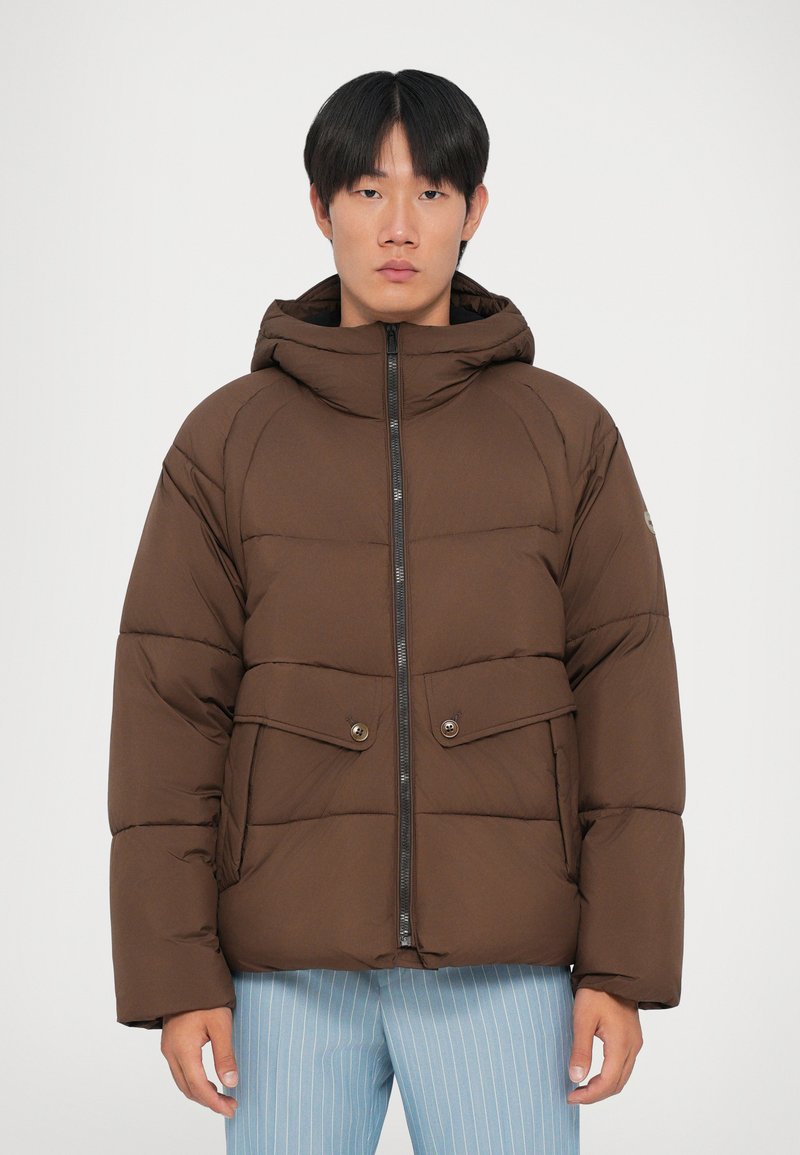Giacca puffer marrone con cappuccio, dotata di una cerniera frontale, due tasche con bottone e una texture trapuntata. Indossata con pantaloni a righe blu chiaro.