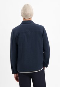Donkerblauwe wollen jas met knoopsluiting bij de manchetten, een kraag en een subtiele textuur. Een beige gebreide beanie maakt de look compleet.