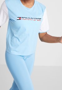 Světle modré tričko s krátkými bílými rukávy, na kterém je červené a námořnické logo "TOMMY SPORT". Vydáváno s odpovídajícími světle modrými legínami.