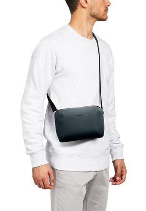 Uomo che indossa una felpa bianca e pantaloni grigio chiaro con una borsa nera a tracolla all'altezza della vita, che guarda verso destra.