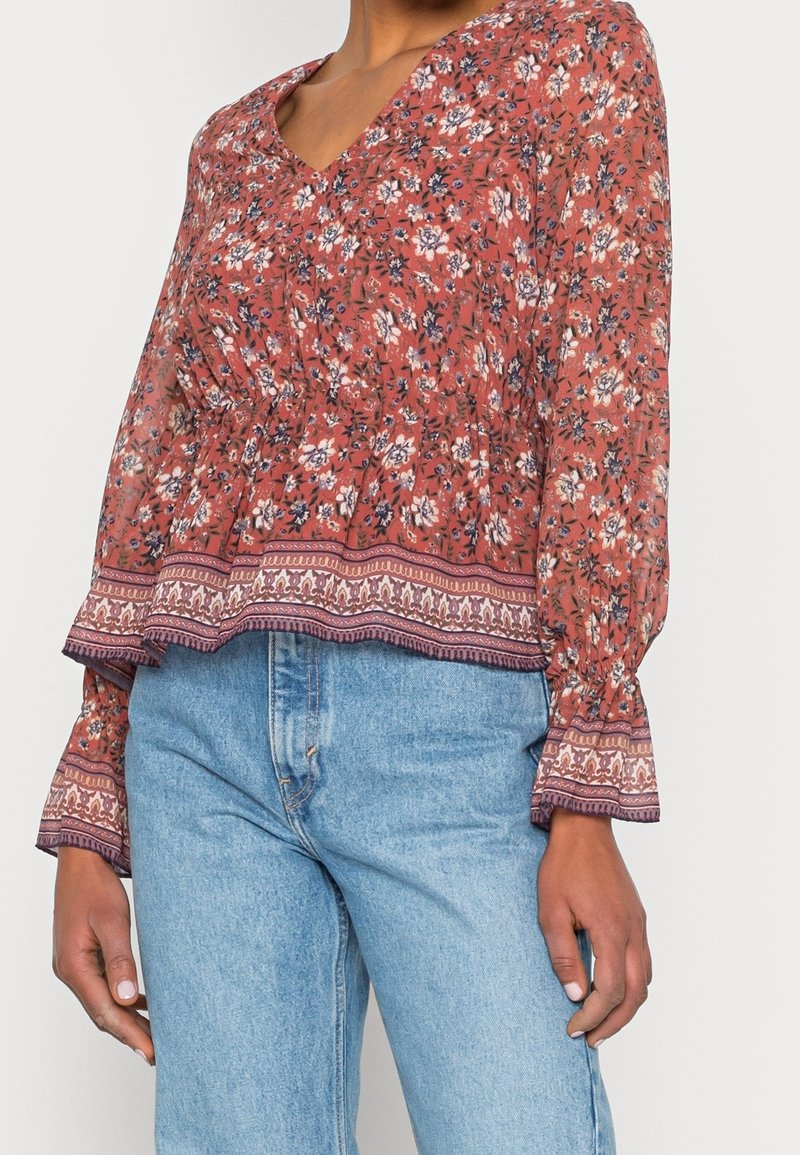 Blusa floreale rossa con maniche lunghe, scollatura a V e orlo arricciato. Tessuto leggero con motivo e una bordatura decorativa in contrasto nella parte inferiore.
