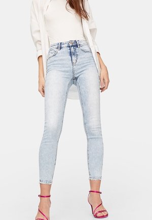 Kobieta w jasnoniebieskich obcisłych jeansach z wysokim stanem, białym topie i otwartej białej koszuli, w połączeniu z jaskraworóżowymi sandałami na obcasie z paskami, stojąca na jednolitym tle.