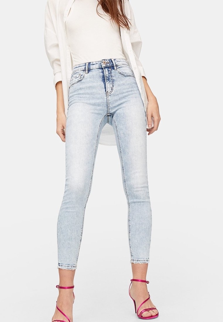 Femme portant un jean skinny taille haute bleu clair, un top blanc et une chemise blanche ouverte, assortis à des talons à brides rose vif, debout devant un fond uni.