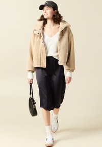 Giacca in pile beige con interno in corduroy, maglione bianco traforato, gonna in satin nera, sneakers bianche e borsa nera.