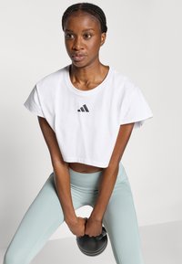 Λευκό κοντό αθλητικό πουκάμισο με κοντά μανίκια, με μαύρο λογότυπο Adidas. Συνδυάζεται με κολάν μέντας ψηλόμεσο και μια αλτήρα kettlebell.