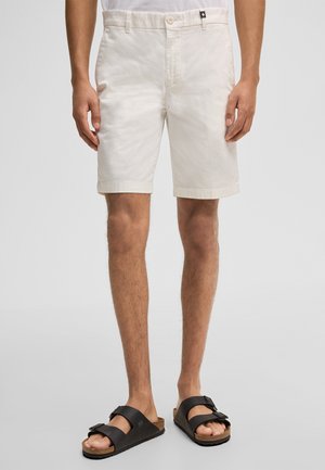 Mannelijk model draagt off-white knielange shorts en zwarte sandalen met dubbele riemen, staand tegen een effen lichtgrijze achtergrond.