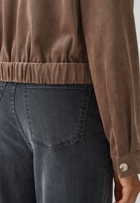 A brown corduroy jacket with a gathered hem and long sleeves, paired with gray denim jeans featuring a back pocket detail.