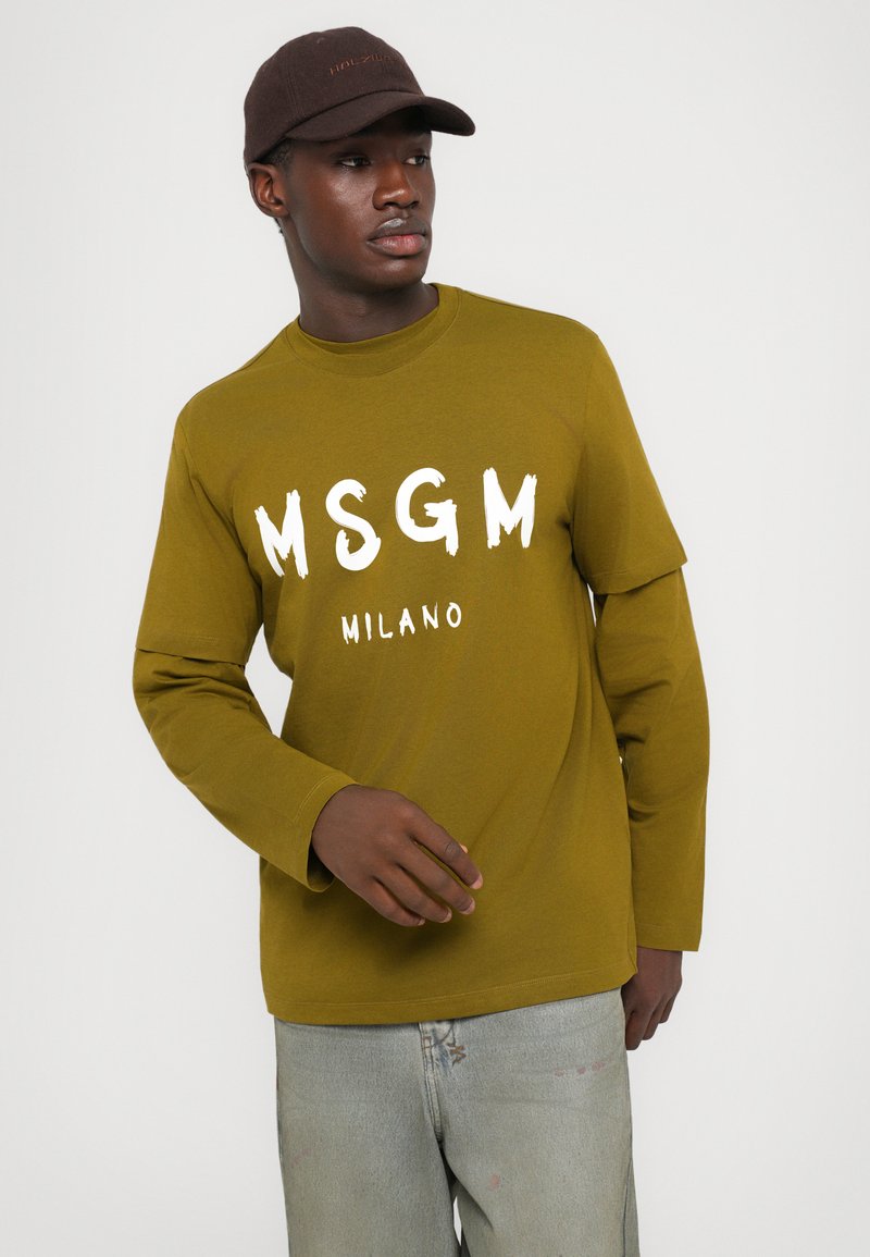 Olivgrön långärmad t-shirt med vit text "MSGM MILANO", kombinerad med ljusblå slitna jeans och en brun keps.