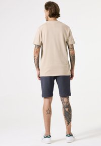 Beige t-shirt, korte mouwen, relaxte pasvorm, gecombineerd met donkergrijze shorts, witte sneakers, zichtbare tatoeages op de benen. Schoon, minimalistisch ontwerp.