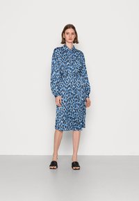 Vestido com padrão de leopardo azul, com gola e mangas longas, ajustado na cintura, apresentando um corte até ao joelho. Usado com sandálias pretas trançadas.