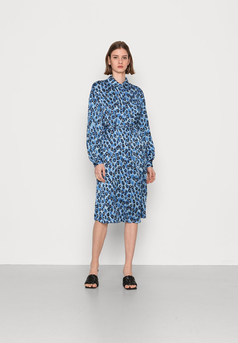 Vestido com padrão de leopardo azul, com gola e mangas longas, ajustado na cintura, apresentando um corte até ao joelho. Usado com sandálias pretas trançadas.