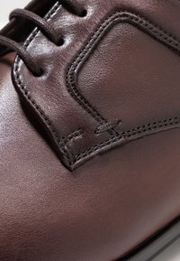 Chaussure en cuir marron avec une texture lisse, des détails cousus et un design à bout arrondi. Les lacets sont marron foncé, se fondant avec la chaussure.