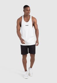 Weißes Tanktop mit dem Aufdruck "SMILODON" auf der Brust, kombiniert mit schwarzen Shorts und weißen Turnschuhen; schlichtes Design, leichtes Material.