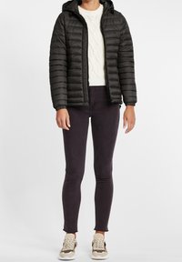 Czarna pikowana kurtka puchowa z kapturem, zestawiona z kremowym swetrem w warkocze, ciemnymi dopasowanymi dżinsami i beżowymi trampkami.