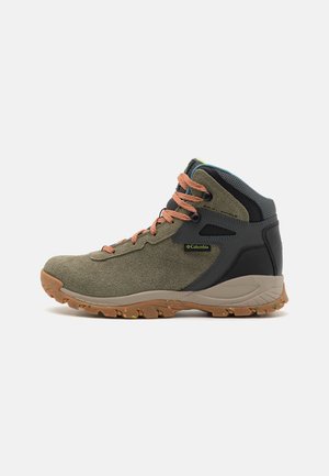 Botas de senderismo Columbia de altura media en ante de color verde olivo, negro y gris, con cordones naranjas y una suela de goma marrón rugosa con motas de colores.