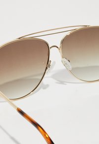 Occhiali da sole aviator con montatura dorata, lenti con gradiente marrone e punte delle tempie a motivo tartaruga su uno sfondo bianco.