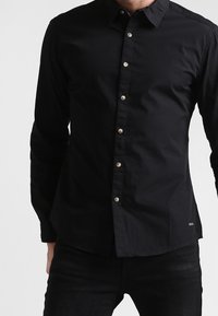 Zwart shirt met lange mouwen gemaakt van een gladde stof; heeft een puntkraag, een knoopsluiting met metalen knopen en een afgeronde zoom.