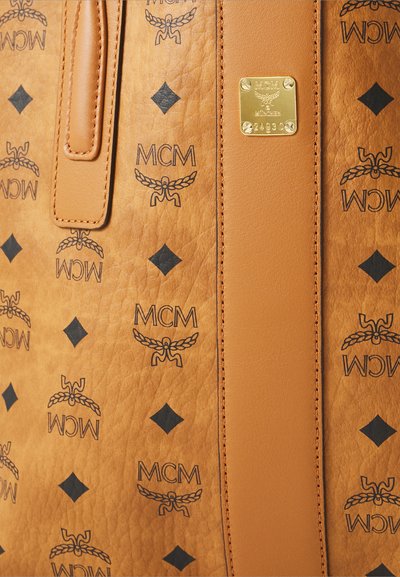 Tan lederen stof met zwart MCM-logo patroon, diamanten vormen en een gouden metalen plaat met merknaam en serienummer.