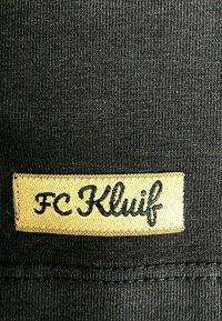 Zwarte ribstof met een rechthoekige gele patch waarop de geborduurde tekst "FC Kluif" in zwart cursief staat.