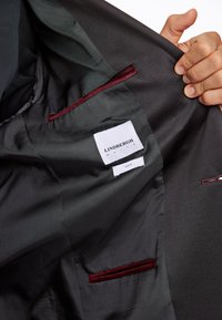 Fekete kabát belseje sima textúrájú, piros bársonyos díszítéssel a zsebeknél és a "LINDBERGH WHITE" és "SLIM FIT" feliratú címkékkel.