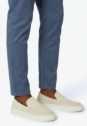 Person wearing blue pants and light beige slip-on sneakers with white soles, standing against a plain light background.