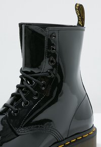 Botte de combat noire brillante à lacets avec piqûres jaunes et languette de tirage marquée sur fond clair.