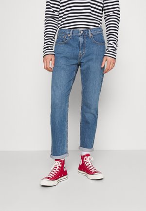 Personne portant un jean bleu retroussé, des baskets montantes rouges et un t-shirt à manches longues rayé noir et blanc, debout sur un sol gris.