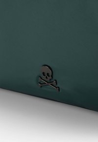 Borsa di tessuto verde scuro con un'emblema lucido di teschio e ossa incrociate come dettaglio distintivo. Texture liscia, forma rettangolare.