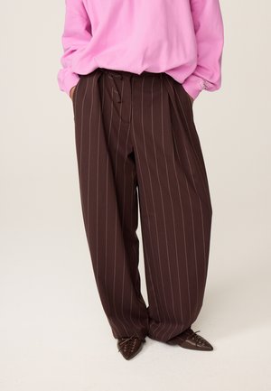 Persoon draagt losse donkerbruine pantalon met krijtstreep, roze top met lange mouwen en bruine veterschoenen met puntige neus, handen in de zakken.
