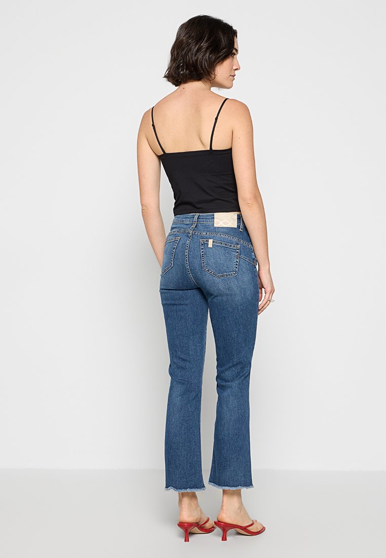 Blaue Jeans im Schlag mit rohem Saum, versehen mit einem beigefarbenen Patch am Bund und zwei Gesäßtaschen. Kombiniert mit einem schwarzen Tanktop.