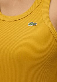 Gros plan d'un débardeur jaune côtelé avec un petit logo crocodile vert brodé sur la poitrine.