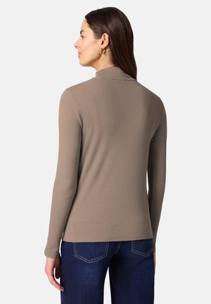 Femme aux cheveux bruns raides portant un haut taupe à manches longues et un jean bleu foncé, tournée vers la gauche sur un fond clair uni.