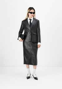 Sort læderblazer med revers og knapper, parret med en matchende blyantnederdel. Hvid skjorte med krave og sort slips. Sorte solbriller og taske.
