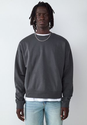 Young man with dark skin and dreadlocks wearing a dark gray sweatshirt, light blue jeans, and a silver chain necklace, standing against a plain background.